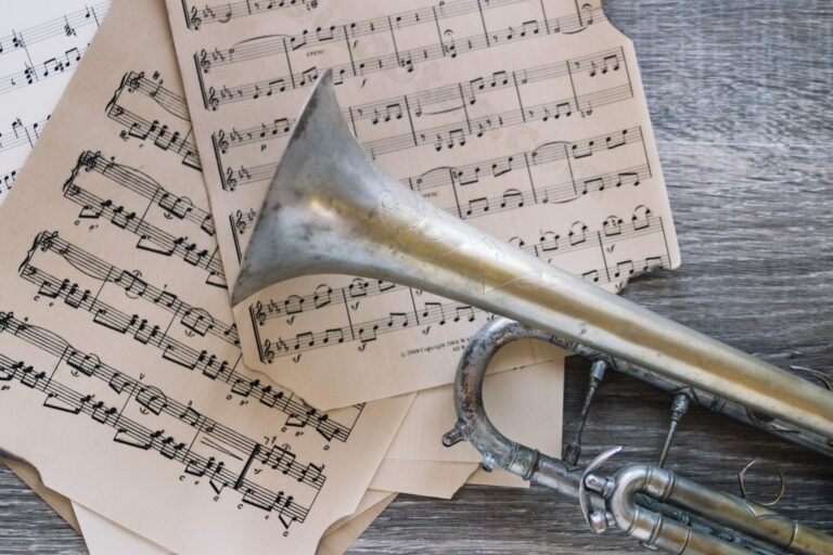 Stara, metalowa trąbka leżąca na drewnianym blacie, ułożona na kilku pożółkłych kartkach z nutami. Instrument ma widoczne ślady zużycia i patynę, a w tle widać zapis nutowy na pięciolinii, co tworzy klimat muzyczny i lekko retro.