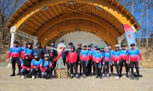 Grupa kilkunastu osób w strojach kolarskich stoi i kuca przed drewnianą zadaszoną sceną w plenerze; w centrum znajduje się kukła przypominająca Marzannę, a uczestnicy mają na sobie kaski i okulary rowerowe.