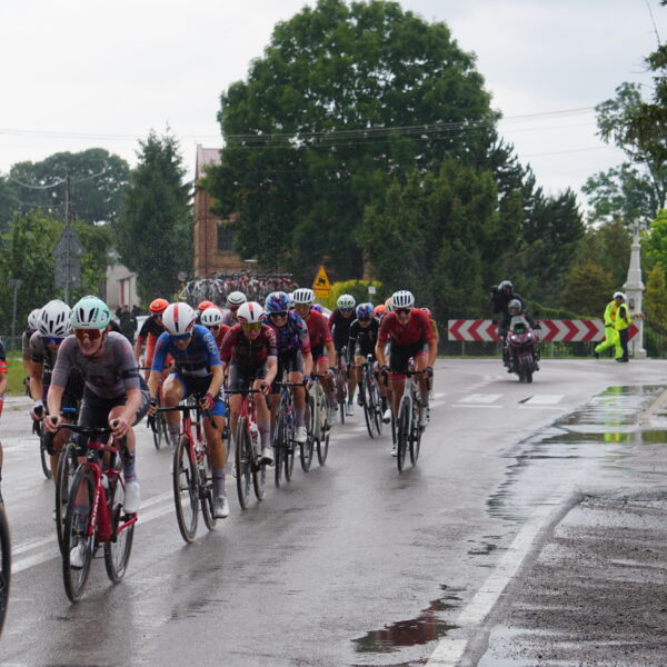 Peleton kolarek uczestniczących w 4. Tour de Pologne Women przejeżdża przez skrzyżowanie w Gorajcu-Starej Wsi na terenie Gminy Radecznica. Przy drodze kibicują mieszkańcy oraz członkowie Brygady RR – Rowerowej Radecznica z flagami i balonem promocyjnym gminy. Nad bezpieczeństwem wyścigu czuwają strażacy i służby porządkowe. Nawierzchnia jezdni jest mokra po opadach deszczu.