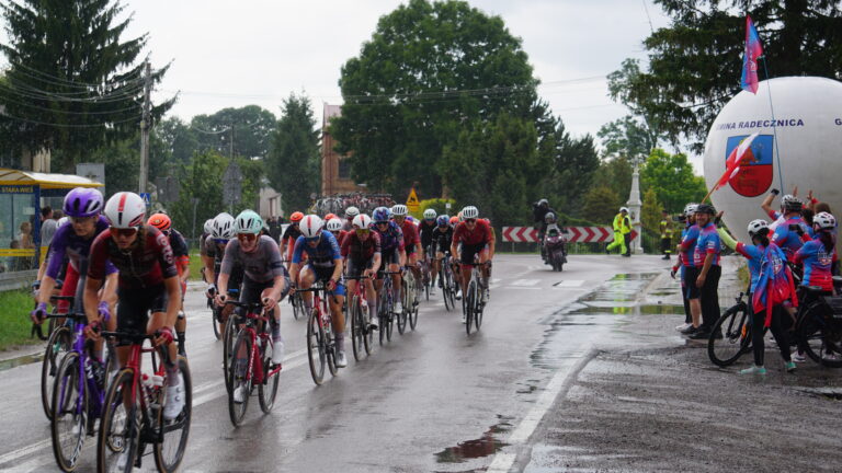 Peleton kolarek uczestniczących w 4. Tour de Pologne Women przejeżdża przez skrzyżowanie w Gorajcu-Starej Wsi na terenie Gminy Radecznica. Przy drodze kibicują mieszkańcy oraz członkowie Brygady RR – Rowerowej Radecznica z flagami i balonem promocyjnym gminy. Nad bezpieczeństwem wyścigu czuwają strażacy i służby porządkowe. Nawierzchnia jezdni jest mokra po opadach deszczu.